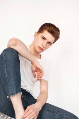 Havalı beyaz tişörtlü, mavi kotlu, şık görünümlü yakışıklı genç adam kapalı alanda klasik duvarın yanında oturuyor. Gençlik kıyafetleri içinde çekici bir adam stüdyoda dinleniyor. Biçim.