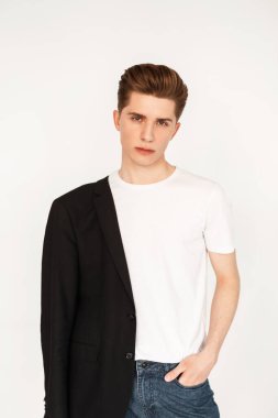 Bir omzunda siyah moda klasik ceketi olan şık beyaz tişörtlü havalı bir genç stüdyoda beyaz arka planda duruyor. Vintage duvarın yanında saç stili olan yakışıklı ciddi adam modeli..