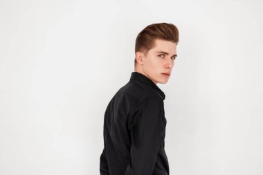 Oldukça ciddi bir portre, kendine güvenen, modaya uygun saç stili, şık siyah gömlekli, klasik beyaz duvarın yanında duran, oldukça ciddi bir genç adam. Çekici, modern model çocuk. Şık giyiniyor.