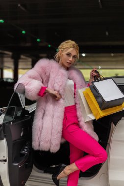 Modern arabanın yanında poz veren şık, pembe kürklü, şık bir manken kadın. Alışveriş poşetleriyle gezintiden sonra dinlenen göz kamaştırıcı bir kız. Bayan alışverişkolik..
