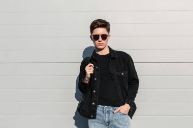 Moda güneş gözlüklü, şık, sıradan kot elbiseli, sırt çantalı modern Amerikalı genç hippi adam, güneşli bir caddede, binanın yakınında dinleniyor. Şehirdeki moda mankeni. Gençlik tarzı.