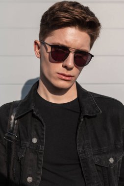 Açık havada güneşli bir günde, duvarın yanında şık güneş gözlükleriyle şık siyah kot ceketli, saç stili havalı genç bir adam. Sokaktaki yakışıklı adam. Sıradan erkek giyimi. Yakından erkek yüzü.