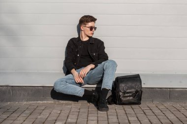 Siyah güneş gözlüklü, şık kot elbiseli, şık spor ayakkabılı, şık deri çantalı Avrupalı genç adam kentin klasik duvarlarının yanındaki güneş ışınlarının keyfini çıkarıyor. Şehir modası hipster çocuğu..