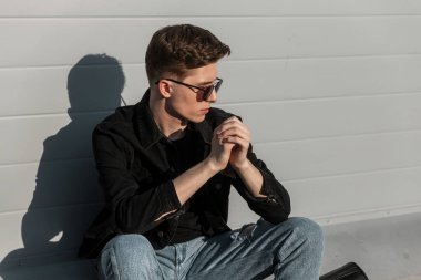 Moda güneş gözlüklü, şık ve günlük kot elbiseli havalı Amerikalı genç hippi, güneşli bir günde apartmanın yakınında dinleniyor. Şehirde modern erkek mankeni. Gençlik tarzı.