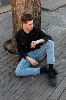 Yakışıklı Amerikalı seksi hippi adam şehrin yazlık terasında ağaçların yanında dinleniyor. Modaya uygun modern bir adam oturur ve dışarı bakar. Erkekler için günlük kot pantolon..