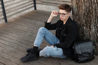 Saç stili, kotlu, kot pantolonlu ve çantalı genç hippi gözlükleri düzeltiyor ve bir ağacın yanına oturuyor.