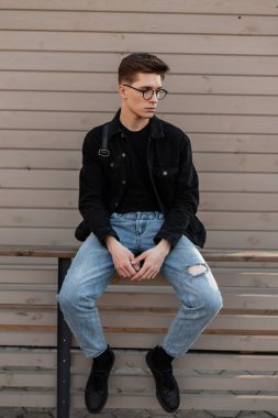 Şık Amerikan erkek hipster, sıradan moda kıyafetleri içinde, deri şık siyah spor ayakkabıları ile kentteki ahşap binanın yanında oturuyor. Modayı takip eden oldukça çekici bir genç sokakta dinleniyor.