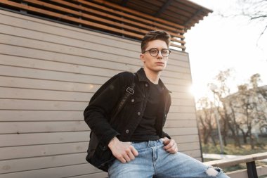 Havalı, modaya uygun kot elbiseli, şık gözlüklü, modaya uygun siyah sırt çantalı Amerikalı havalı genç hippi dışarıda ahşap duvarın yanında dinlenmenin ve güneş ışınlarının keyfini çıkarıyor. Moda adamı ve parlak günbatımı.