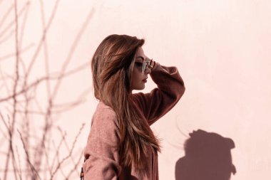 Güzel genç bir kadın şehirde şık saçlarını düzleştiriyor. Moda güneş gözlüklü çekici modern kız modeli açık havada güneşli bir günde pembe duvarın yanında şık bir ceket giyiyor. Profil fotoğrafı.