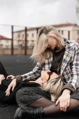 Moda modası geçmiş eski moda kıyafetler içinde güzel kız kardeşler oturur ve birbirlerine bakarlar. Şık ceketli güzel kızlar basketbol sahasında dinleniyor. Kadınlar için moda koleksiyon kıyafeti.