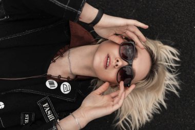 Modern sarışın, şık siyah elbiseli, gözlüklü, açık hava gözlüklü bir kadın. Genç elbiseli seksi kız manken yalan söylüyor ve şehirde asfalta eski model güneş gözlüğü takıyor..