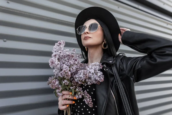 サングラスのスタイリッシュなレザージャケットでファッショナブルな帽子のかなり都会の女性は手にライラックの花束を保持しています 美しい花を屋外でポーズ黒いファッションの服を着た素敵な女の子 — ストック写真