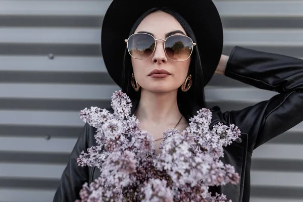 时尚可爱的女人 戴着时髦的太阳镜 头戴老式的帽子 穿着时髦的皮夹克 头戴紫丁香花束 在这座城市的金属建筑附近摆设 迷人的女孩在城市里走来走去 — 图库照片