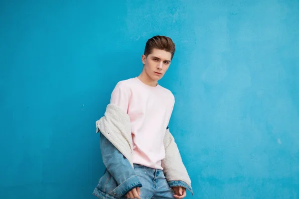 Attractive young man with hairstyle in stylish pink sweatshirt in fashionable denim jacket posing near vintage colored wall in street. European handsome guy in jeans clothes is stand blue background.