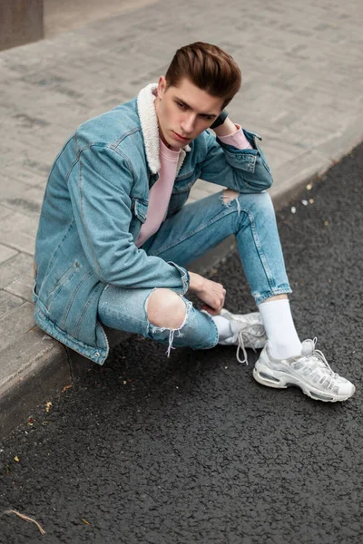 Moda mankeni, şık kot ceketli, modaya uygun kot pantolonlu, şık beyaz ayakkabılı genç adam şehrin yol kenarında kaldırımda oturuyor. Sokakta günlük giysiler içinde yakışıklı bir adam..