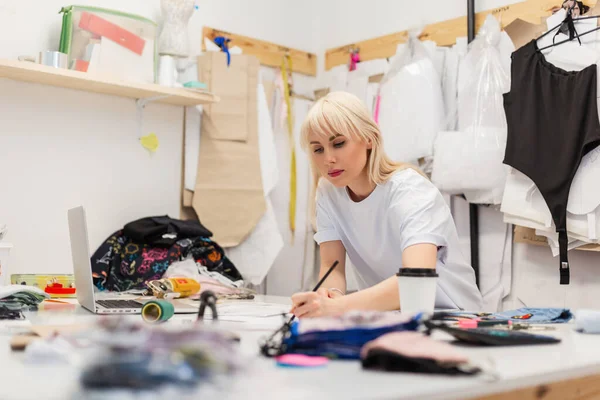 Young beautiful woman fashion designer working in atelier. Workspace of a creative fashion designer with a laptop, coffee, scissors, threads, fabrics and other elements