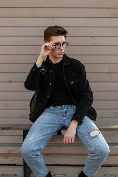 スタイリッシュなジーンズの服を着たファッションモデルのクールな若い男は流行のメガネをまっすぐにします ストリートの木製の壁の近くに黒い革のバックパックのポーズを持つカジュアルなデニムウェアのアメリカのスタイリッシュな男 青年服 — ストック写真