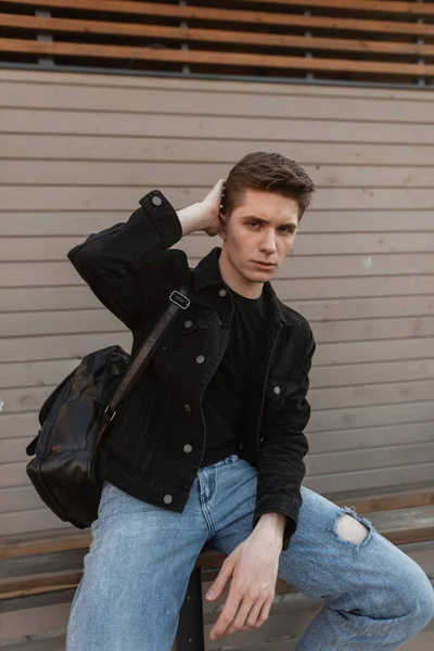 スタイリッシュなジーンズの服のかなり魅力的な若い男は流行の髪型をまっすぐにします ストリートの木製の壁の近くに休んで黒革のバックパックとカジュアルなデニムウェアでスタイリッシュなハンサムな男 ユース スタイル — ストック写真