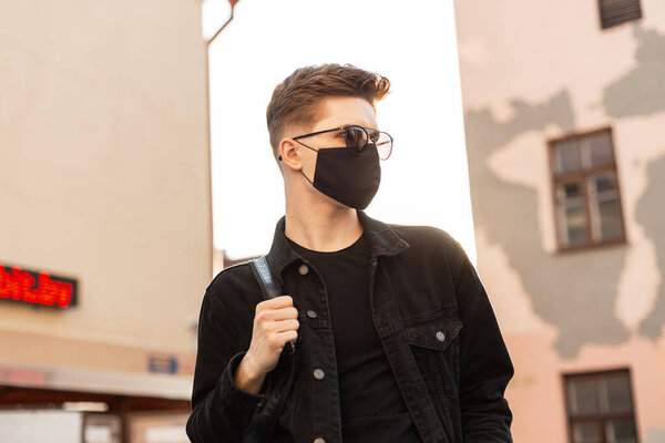 Young man in trendy sunglasses in denim jacket in protective black mask stands on city and enjoys the bright spring day. Fashionable teenage guy in mask is protected from Covid-19. Healthy lifestyle.