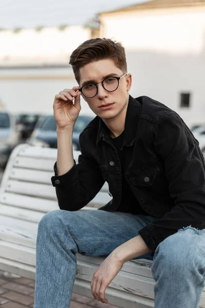 スタイリッシュなジーンズの服を着たファッションモデルのクールな若い男は流行のメガネをまっすぐにします ストリートのベンチにブラックレザーバックパックポーズでカジュアルなデニムウェアでアメリカのスタイリッシュな男 若者服 メンズウェア — ストック写真