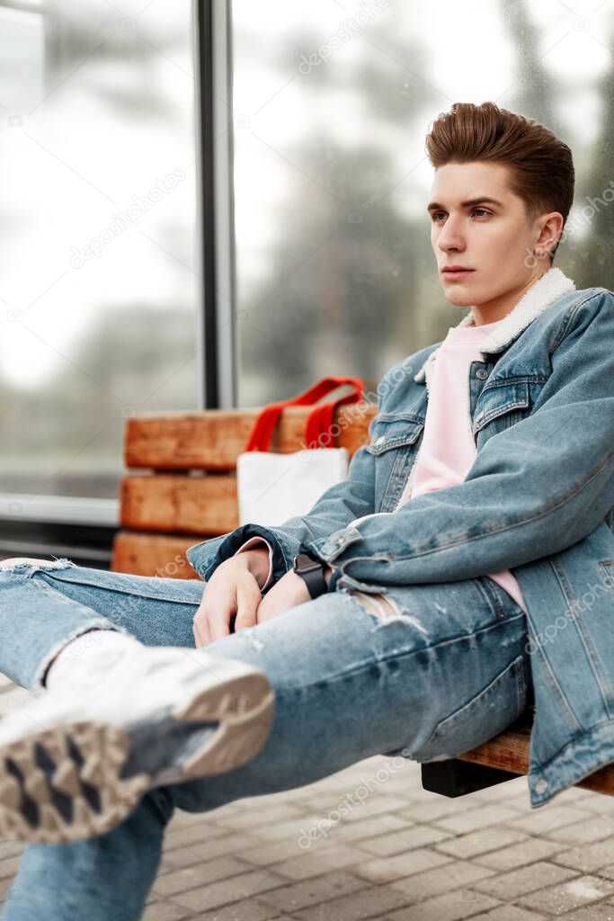 Trendy young man in youth stylish denim jacket in casual vintage jeans with fabric shopper sit on wooden bench at stop of public building in city. Good-looking guy in fashionable clothes on street.