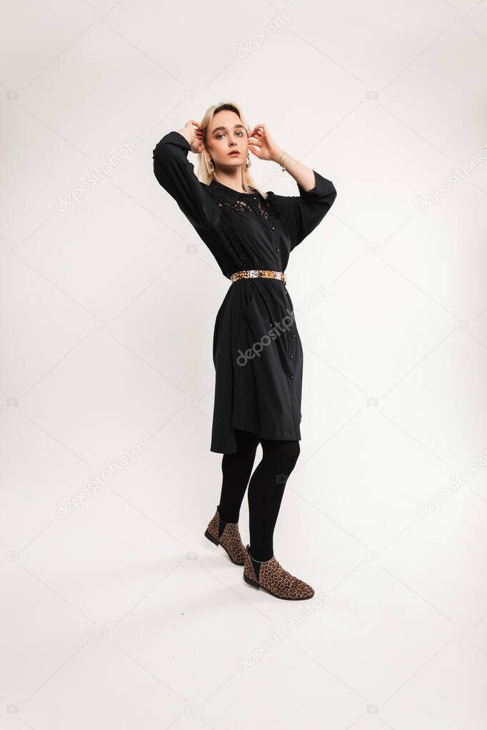 Elegant lovely young woman blonde in black dress with leopard strap in trendy leopard shoes stands and straightens hairstyle in studio. Pretty fine stylish girl fashion model in retro outfit indoors.