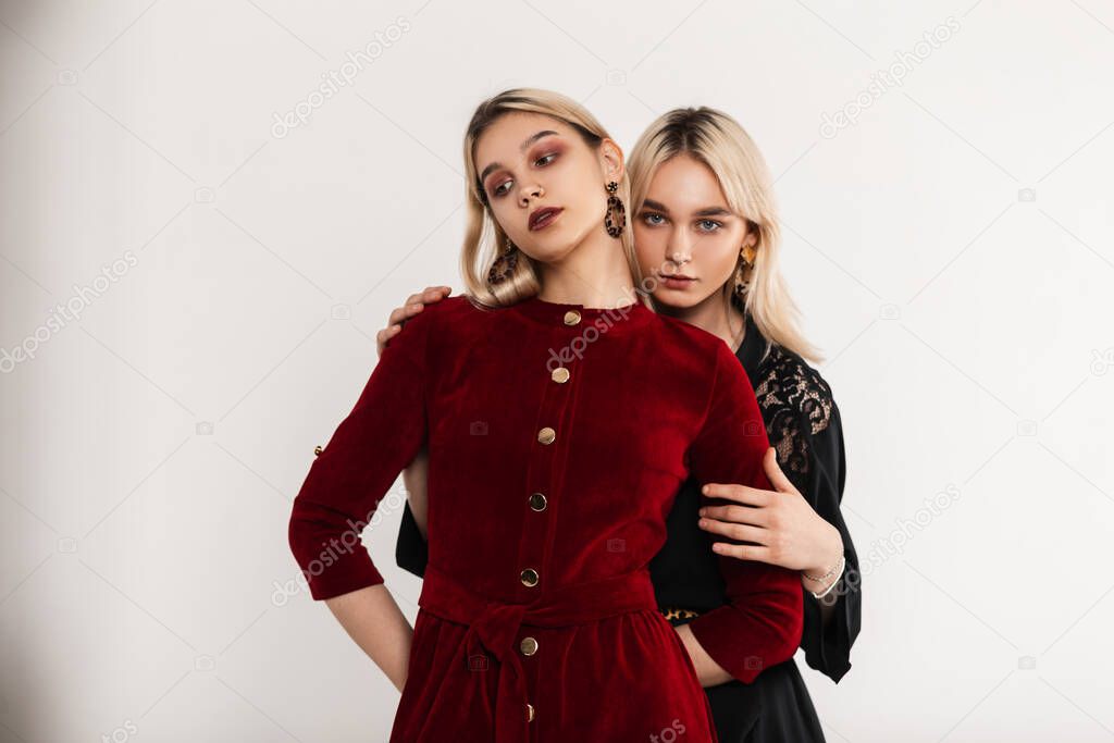 Two pretty young girlfriend blondes in stylish dresses with sexy lips posing near white vintage wall indoors. Lovely gorgeous modern beautiful lesbian girls in fashionable outfit in studio. Fashion.