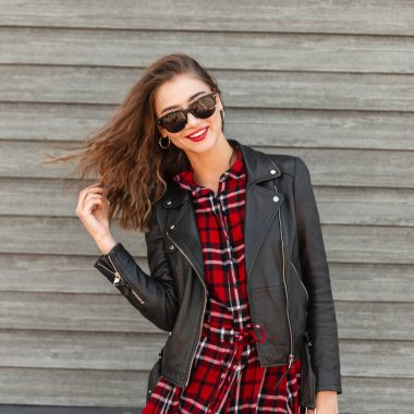 Güzel, güzel, neşeli, tatlı gülüşü olan, moda gençliğinde güneş gözlüklü, kırmızı-siyah elbiseli, yaz gününde tahta vintage duvarın yanında poz veren genç bir kadın. Güzel, mutlu, gülümseyen kız şehirde yürüyor..