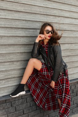 Kırmızı rujlu, güneş gözlüklü, genç, siyah deri ceketli, güzel bir kadın. Ayakkabılı, tahta duvarın yanında duran kırmızı, kareli bir elbise. Sokaktaki modern, çekici moda kadını.