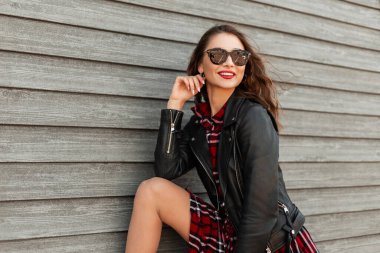 Kırmızı dudaklı mutlu bir kadın, şık güneş gözlüklü, kırmızı kareli elbiseli, siyah deri ceketli, yaz aylarında tahta duvarın yanında poz veren sevimli bir gülümseme. Güzel gülümseyen modern kadın