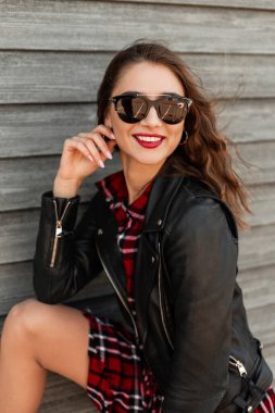 Kırmızı dudaklı, güneş gözlüklü, siyah deri ceketli, kırmızı kareli elbiseli güzel bir kadın yazın sokağın duvarının yanında durur. Mutlu gülümseyen stil kızı dışarıda