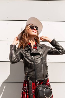 Güneş gözlüklü, siyah deri ceketli, damalı kırmızı elbiseli, büyüleyici bir gülümsemesi olan modern mutlu genç kadın güneşli bir yaz gününde güneşlenmeyi sever. Güzel moda kadını dışarıda güneşlenmeyi sever..