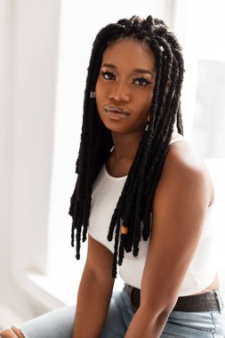 Beyaz tişörtlü, rastalı saç modelli siyah bir kız beyaz bir duvarın yanında oturuyor.