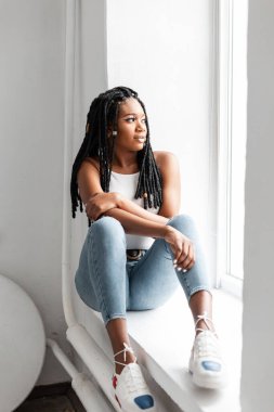 Havalı, modaya uygun, spor ayakkabılı, sıradan kot elbiseli, şık rastalı güzel Afro-Amerikalı kadın içeride pencereye oturuyor. Çekici trend siyah kız modeli beyaz stüdyoda oturuyor..
