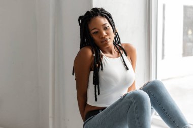 Moda gençliği kıyafetleri içinde şık siyah saçlı modern havalı Amerikan model kadın aydınlık bir günde pencerenin yanında oturur. Çekici siyah kız evde rahatlıyor..