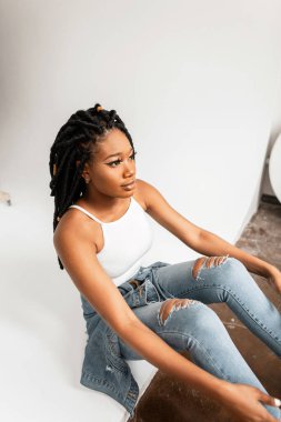 Kotlu, şık, mavi elbiseli, rastalı giysili moda Afrikalı Amerikalı kadın kapalı alanda beyaz duvarın yanında oturuyor. Gençliğinde seksi dudakları olan şık siyahi kız yerde oturuyor.