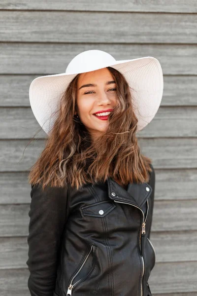 街の木製の壁の近くに黒のスタイリッシュなジャケットで美しいスタイリッシュな帽子で楽しい笑顔で肯定的な若い魅力的な女性モデル 笑顔幸せな女の子とともに赤い唇でカジュアル服屋外 — ストック写真