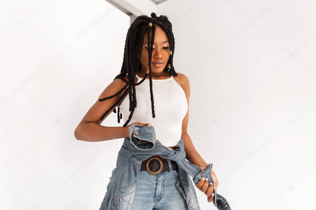 Fashionable afro woman with cool dreadlocks with white top in stylish jeans in trendy denim jacket at waist. Beautiful african american woman with fashion hairstyle in vintage clothes indoors