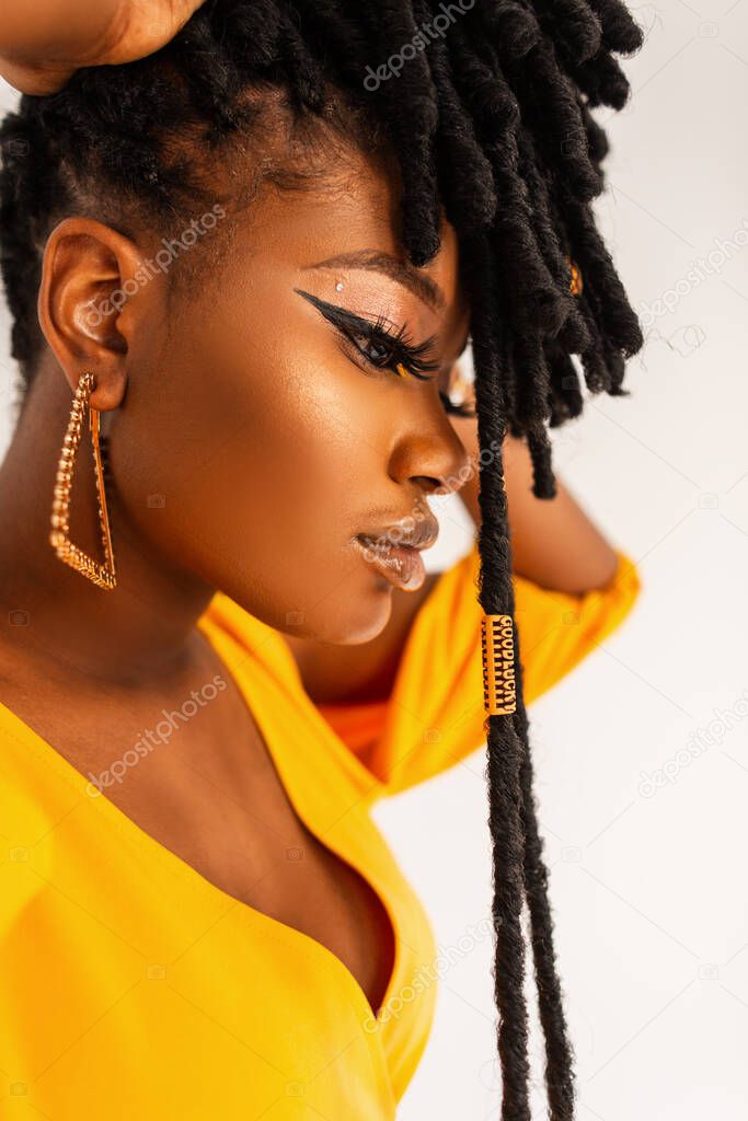 Fashion portrait of model beautiful African woman with clean healthy skin with with gold jewelry earrings with fashionable hairstyle dreadlocks in yellow dress on white background in studio.