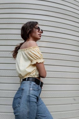 Modaya uygun güneş gözlüklü, mavi, yüksek bel kotlu havalı hippi kadın modeli plastik kaplama duvarın yanında duruyor.