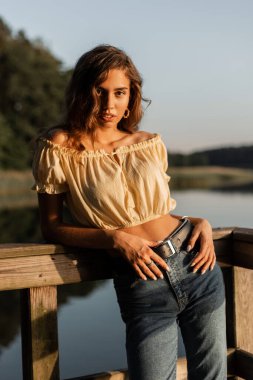 Sarı tişörtlü ve şık kot pantolonlu güzel bir kadın göl kenarında duruyor.