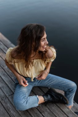 Komik kıvırcık saçlı, neşeli duyguları olan, kotlu, bluzlu ve botlu, şık yaz kıyafetleriyle gülümseyen güzel bir manken kadın denizin kıyısındaki ahşap bir iskelede oturuyor ve anın tadını çıkarıyor.