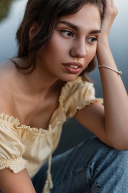 Havalı, sarı bluzlu ve kot pantolonlu çekici, kıvırcık bir kadının yaz portresi doğada göl kenarında oturuyor..