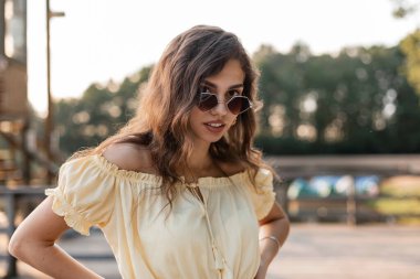 Moda gözlüklü, sarı yazlık elbiseli güzel, kıvırcık bir hippinin taze portresi gölün kıyısında duruyor.