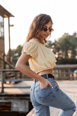 Güzel güneş gözlüklü, şık mavi kotlu ve sarı bluzlu mutlu yaz hanımı gün batımında doğada yürüyor.