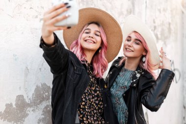 Pembe saçlı, saman gibi şık şapkalı iki genç kadın sokaktaki beyaz duvarın yanındaki modern fotoğraf makinesinde selfie çekiyor. Mutlu kızlar gülümsüyor ve resim çekiyorlar..
