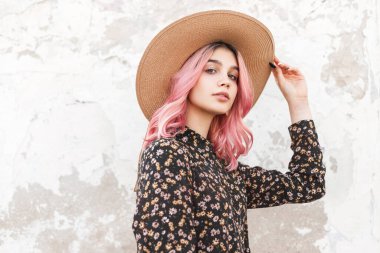 Moda modası geçmiş hasır şapkalı, caddenin yakınındaki güzel çiçek desenli elbisesiyle şık bir genç kadının güzel portresi. Cazibeli, pembe saçlı güzel kız modeli açık havada şapkasını düzeltiyor.
