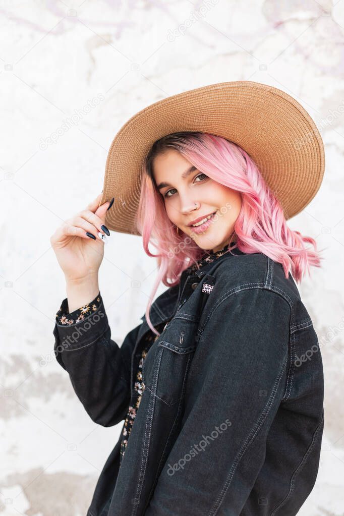 Portrait happy young woman with cute smile with glamour pink hair in stylish denim casual jacket near wall on street. Smiling girl model straightens fashion vintage straw hat outdoors. Summer style.