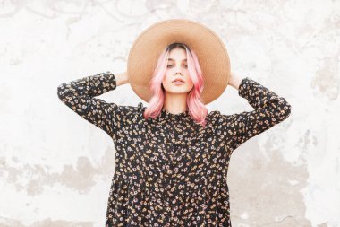 Güzel, pembe saçlı, seksi dudaklı, klasik hasır şapkalı zarif, siyah, şık elbiseli, kentin duvarında poz veren güzel bir kadın. Çekici kız modeli şehirde dinlenmeyi seviyor. Gençlik tarzı.