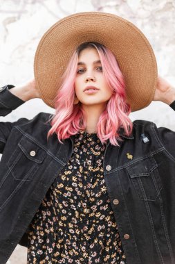 Güzellik portresi göz kamaştırıcı pembe saçlı, şık kot ceketli, sokakta duvarın yanında genç bir kadın. Moda mankeni, açık havada hasır şapka düzeltiyor. Yaz stili.